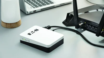 Das Bild zeigt eine liegende Eaton 3S Mini USV, einen Internet-Router, eine kleine Tischlampe und einen Laptop auf einer mittelgrauen Tischplatte. Die Eaton 3S Mini USV ist angeschlossen an den Internet-Router.