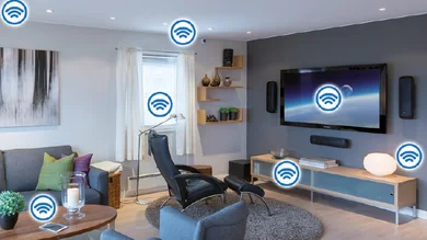 Das Bild zeigt einen gemütlichen Wohnraum mit einem Sofa. An einer Wand ist ein großer Bildschirm befestigt mit Lautsprechern rechts und links Darunter steht eine Konsole mit einer Lampe. Vor der Konsole steht ein Stuhl mit Armlehnen. Rechts und links vom Sofa stehen Stehleuchten. Im Bild sind Symbole für drahtlose Kommunikation bei den Lampen , dem Bildschirm und einem Wandschalter angebracht.